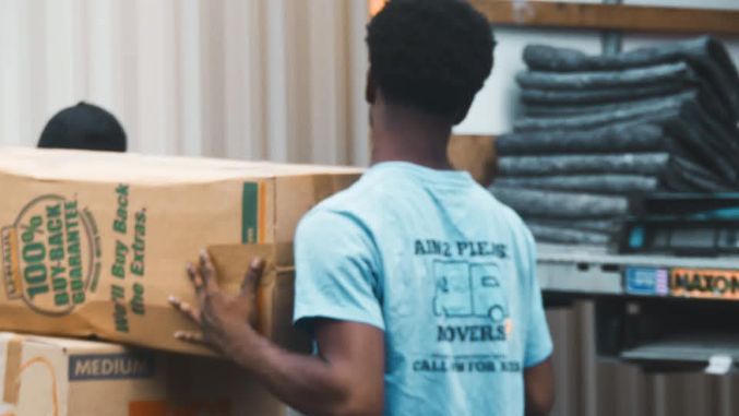 Man carrying large cardboard box in warehouse with stacked materials