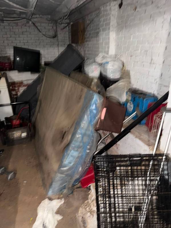 Cluttered basement room with stacked mattresses, boxes, and household items against white brick walls