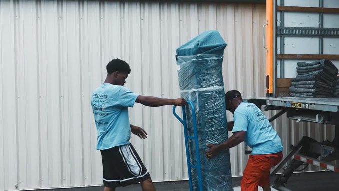 Two workers in blue uniforms loading wrapped furniture onto a truck